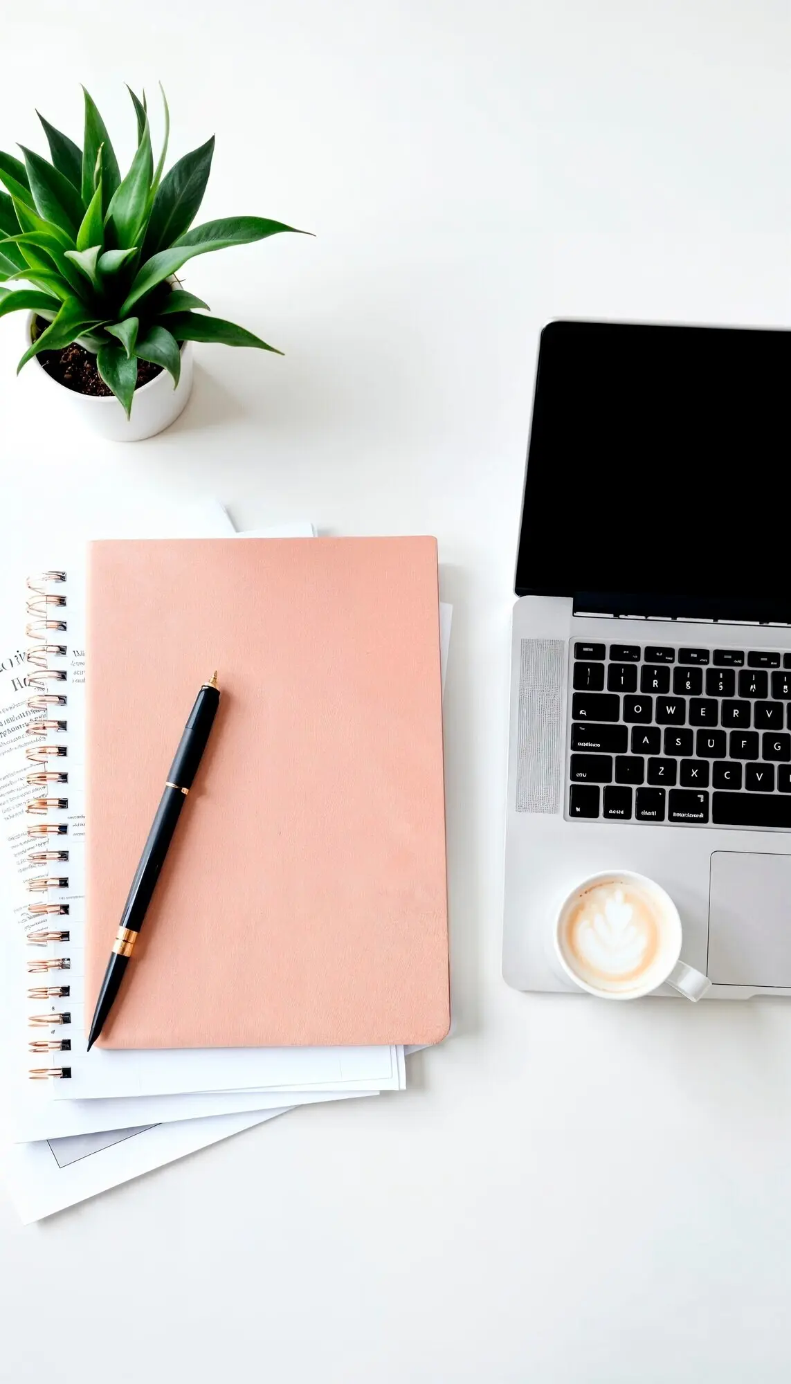 Ein Notizbuch mit einem darauf liegenden Stift neben einem Laptop und einer Tasse Kaffee.